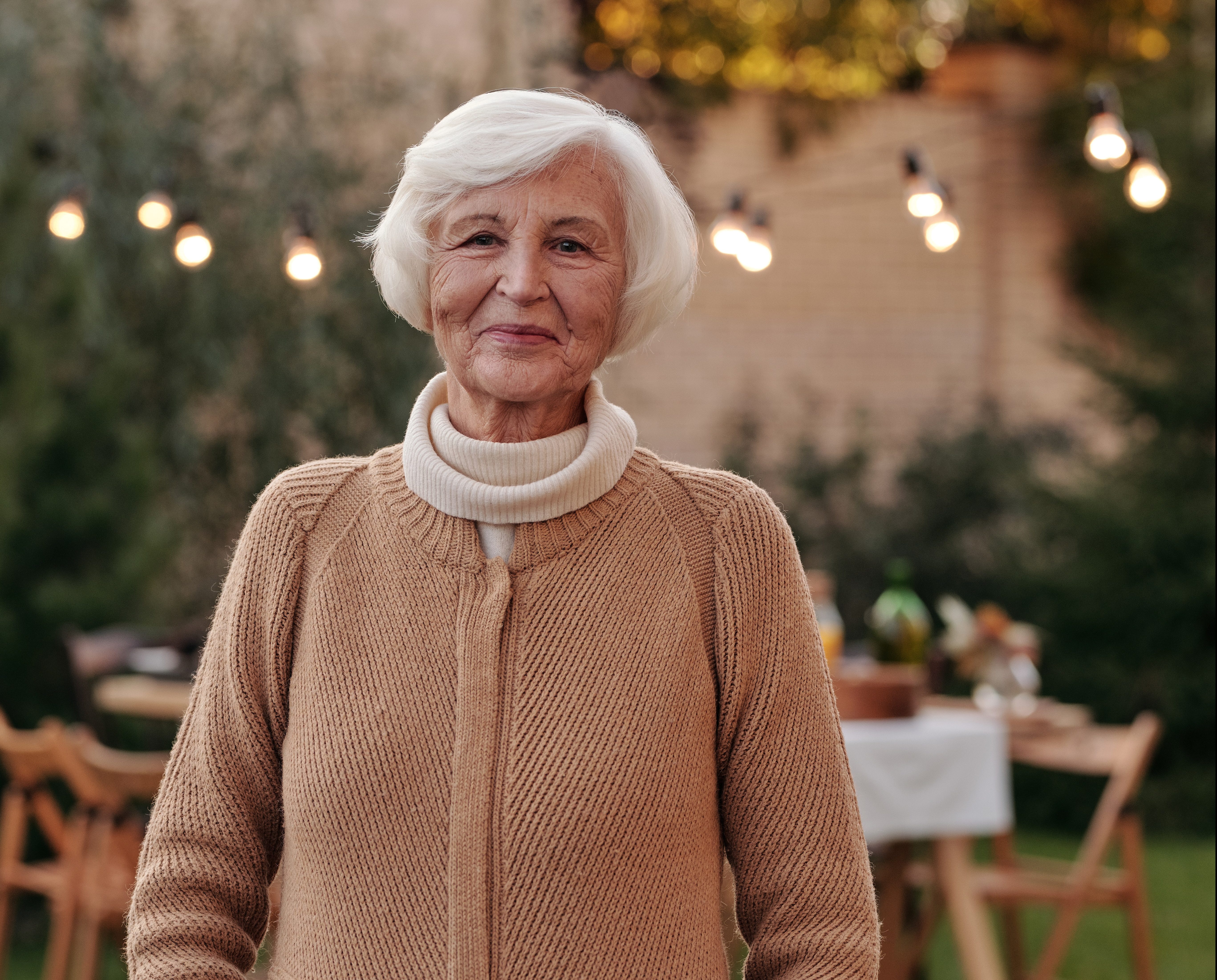Elderly woman with white hair standing in front of an outdoor picnic table with fair lights smiling at the camera