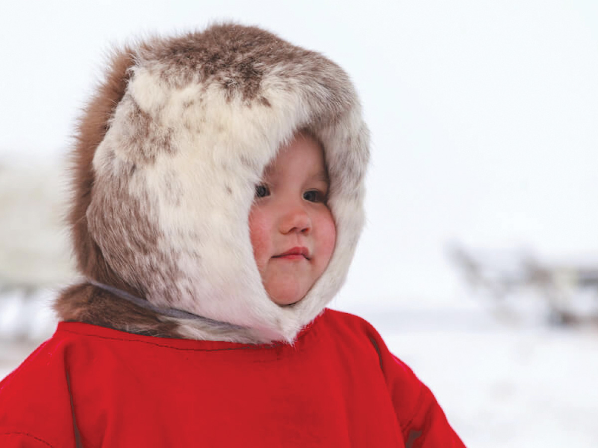 A young boy stands in a traditional Inuit winter outfit including a full fur hood.