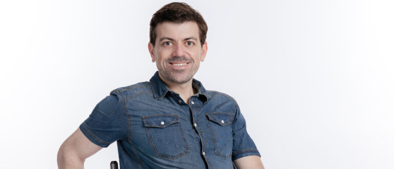 a man in a wheelchair with dark hair, dark eyes, and wearing a denim shirt smiles at the camera