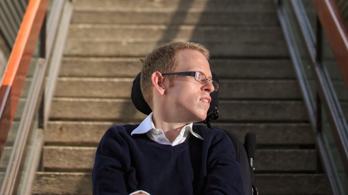 A man in a wheelchair sits at the base of a large set of stairs looking away from the camera.
