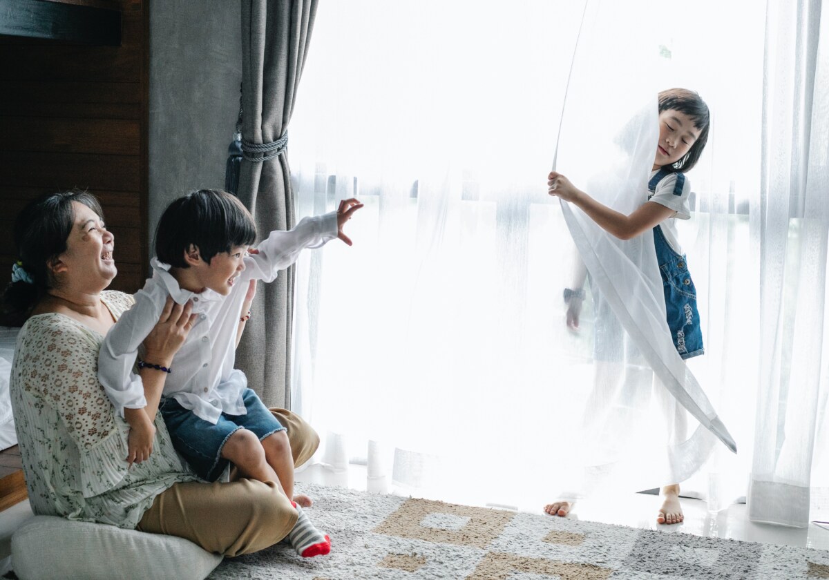 A mother playing with her son while her daughter hides being a curtain laughing