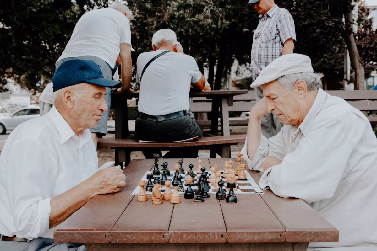 Two older men play chess in a park while a group of men play chess behind them.