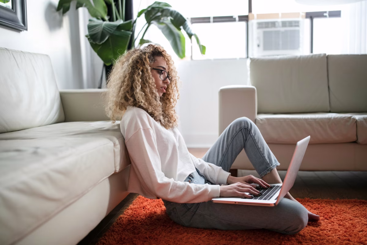 A woman sits on the floor against a couch working on a laptop.