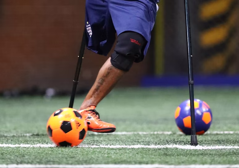 the lower half of a man is visible on a soccer pitch with two soccer balls. the man is missing his right leg and is using 2 crutches for stability.