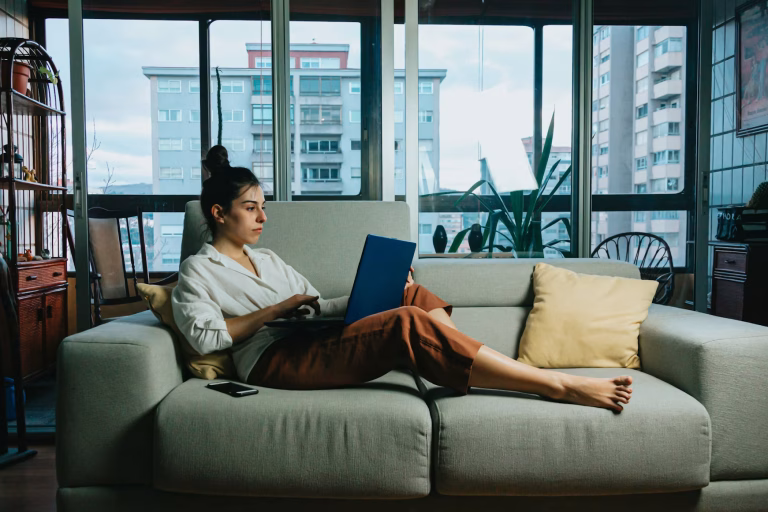 A woman sits across a loveseat with her legs out working on a laptop with a phone beside her.