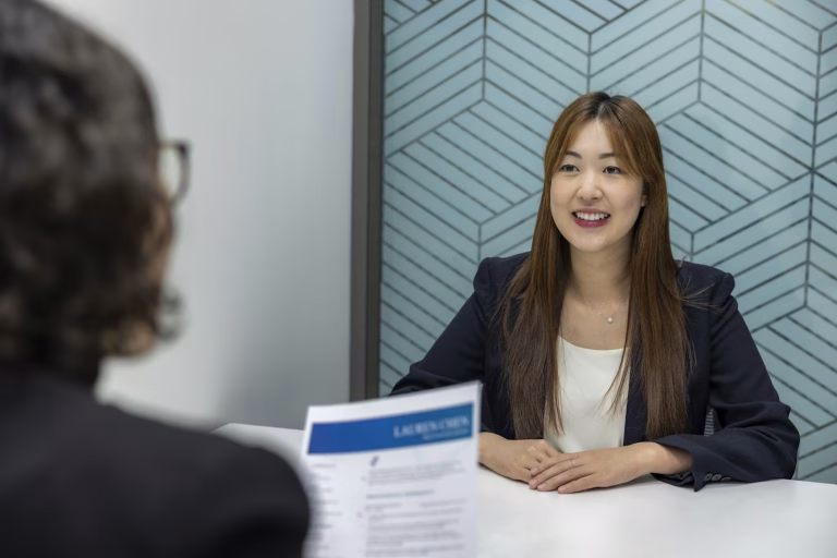 A young woman in business attire sits at a desk facing another woman who is holding a printed resume.