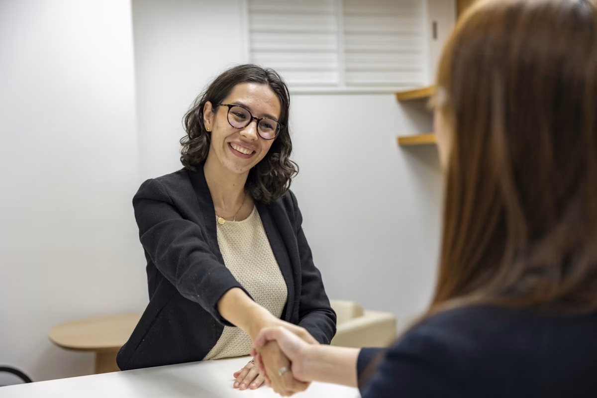 Two women shake hands over a table during a job interview.