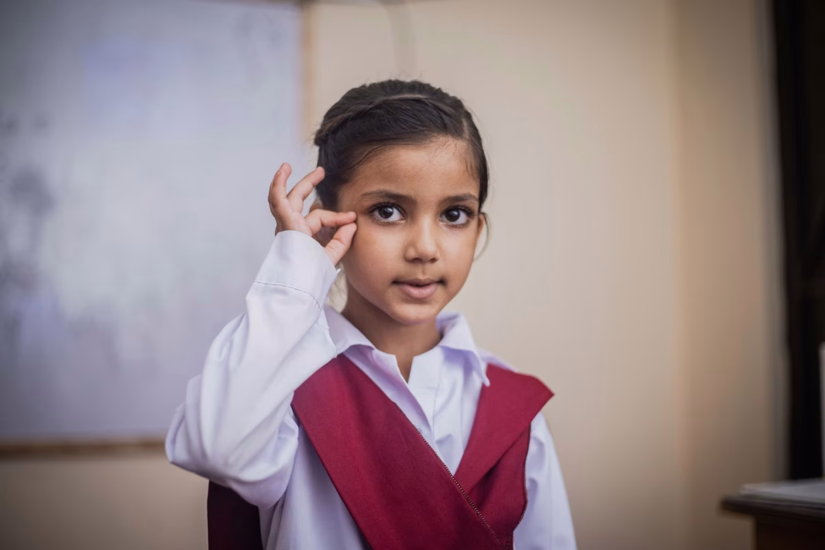 A young girl with dark hair and eyes makes a sign with her hand at her eye and her thumb and forefinger closed and the rest of her fingers straight up.